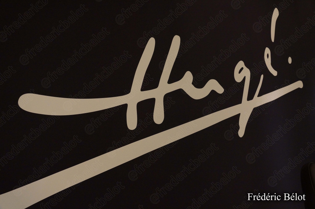 La signature manuscrite d'Hergé agrandie sur un mur sombre de l'exposition.