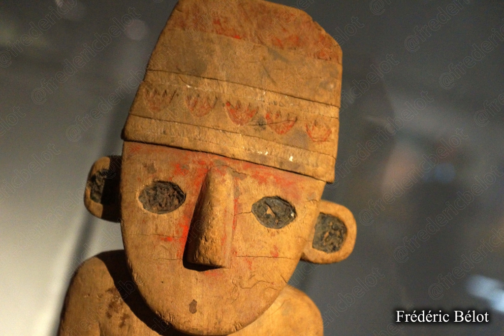 Une sculpture en bois représentant une tête stylisée, coiffée d'un couvre-chef rouge.