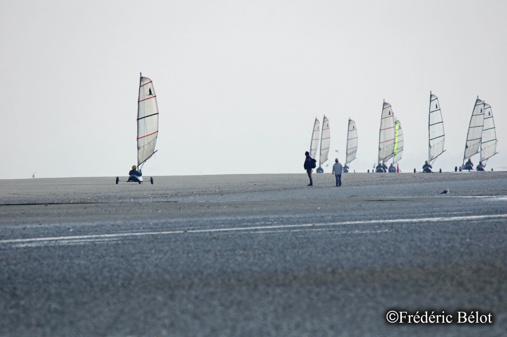 Une série de chars à voile colorés glisse sur le sable, silhouettes de pilotes visibles.