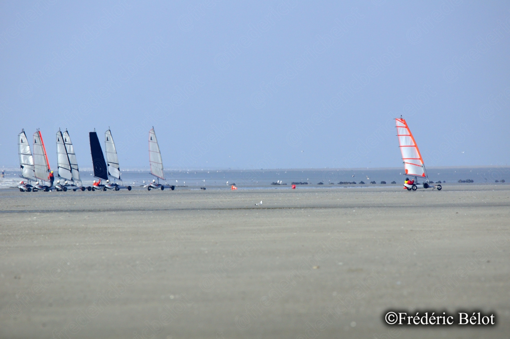 Des chars à voile aux couleurs vives filent sur l'estran, vus de loin.