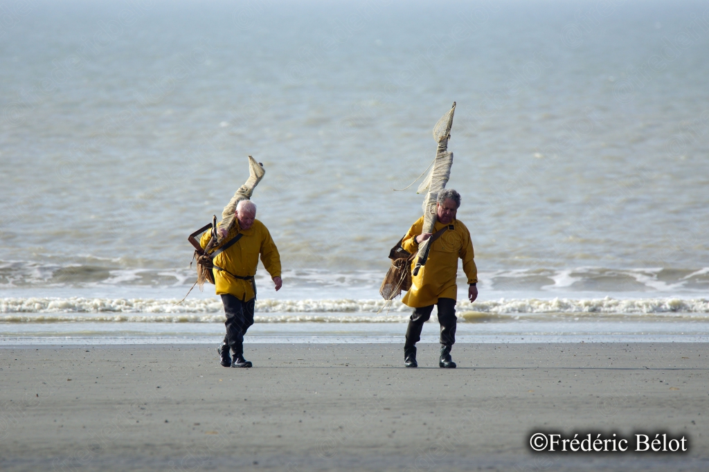 Deux pêcheurs à pied en ciré jaune portent leur haveneau sur l'épaule en remontant de la plage.