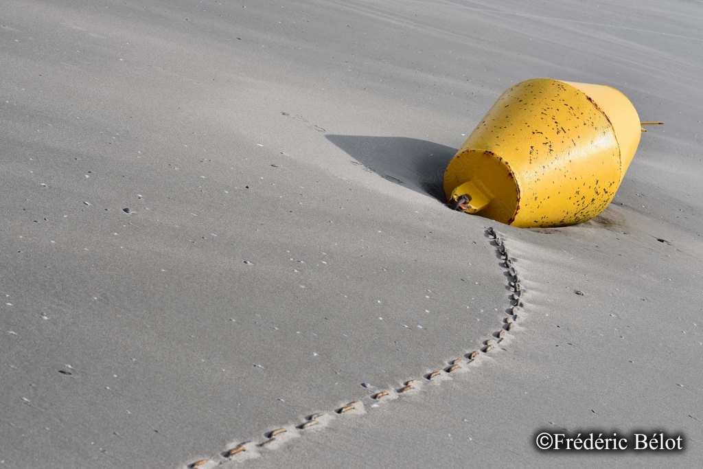 Une bouée jaune échouée sur le sable, avec une longue trace laissée par le vent ou une marée.