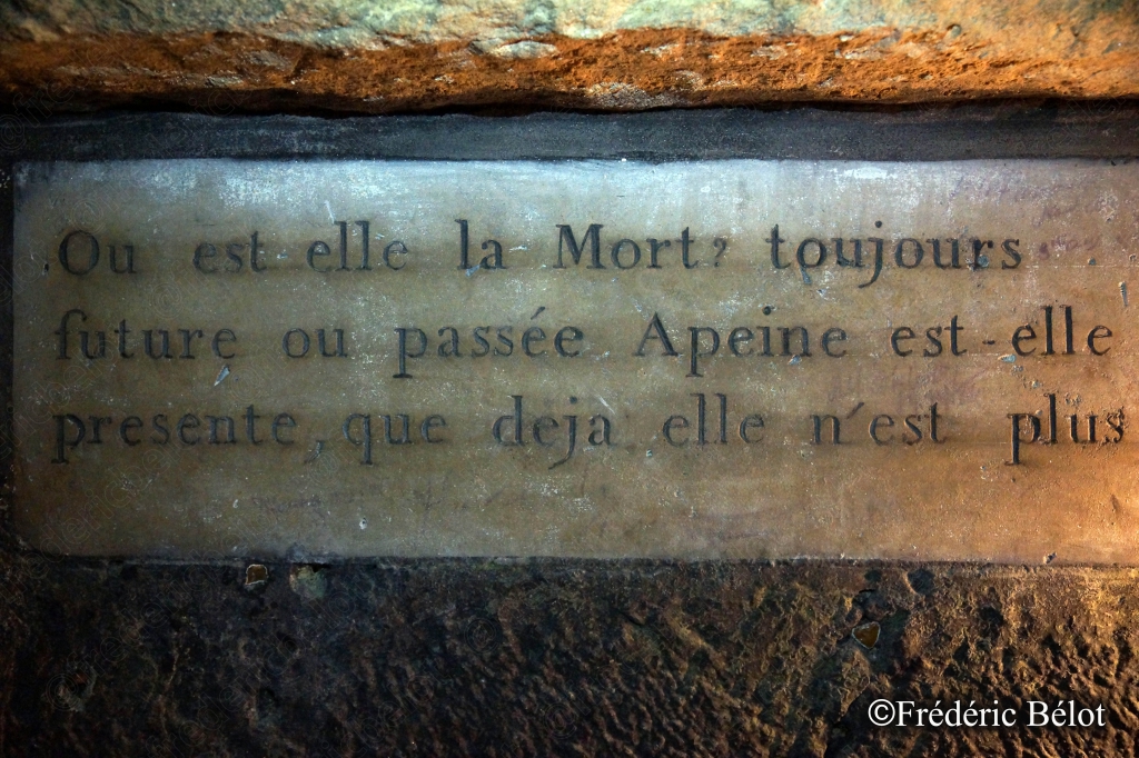 Une inscription gravée dans la pierre évoque la fugacité de la mort, citation posée sur un muret d'ossements.